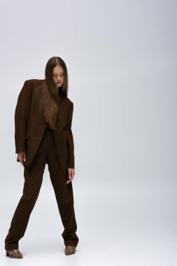full length of brunette teenage model in brown high-quality suit posing on grey