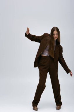 full length of stylish teenage model in brown high-quality suit gesturing on grey