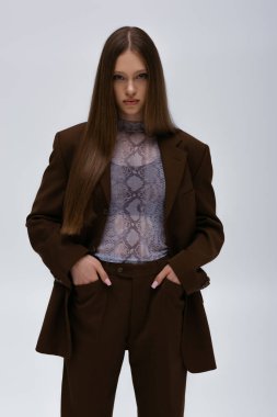 pretty teenage model in brown high-quality suit posing with hands in pockets isolated on grey