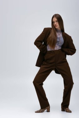 full length of trendy teenage model in brown high-quality suit posing on grey