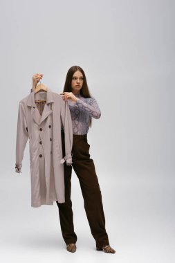 full length of teenage model holding recyclable trench coat on hanger on grey