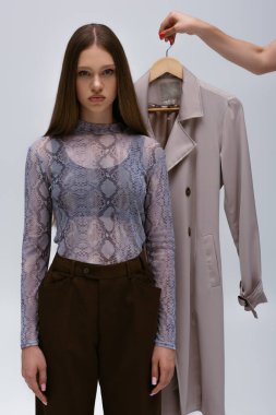 teenage model near woman holding recyclable trench coat on hanger isolated on grey