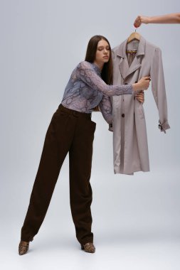 full length of teenage model hugging stylish trench coat while woman holding hanger on grey