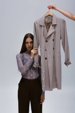 teenage model touching sleeve of recyclable trench coat while woman holding hanger isolated on grey