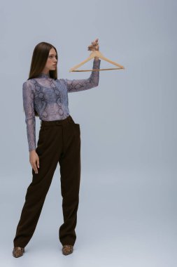 full length of brunette teenage model posing with wooden hanger on grey