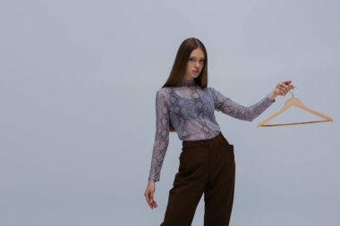 brunette teenage girl holding wooden hanger while posing isolated on grey