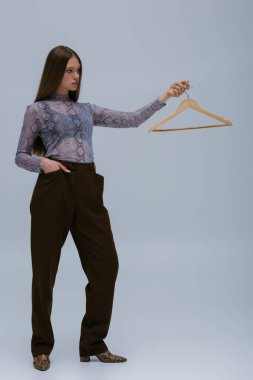 brunette teenage model looking at wooden hanger while posing with hand in pocket on grey