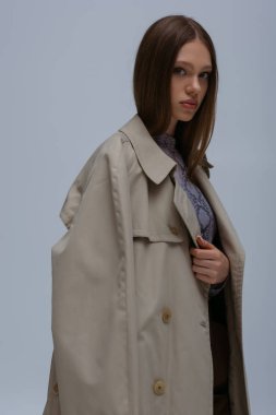 teenage model posing in stylish trench coat and looking at camera isolated on grey 