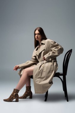 full length of brunette model in trench coat sitting on chair and posing on grey