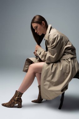 full length of model in stylish trench coat sitting on chair and posing on grey