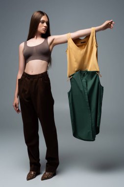 full length of brunette model in crop top and trousers standing with outstretched hand and holding clothing on grey