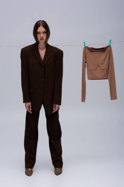 full length of brunette model in brown suit posing near long sleeve top hanging with clothespins on grey 