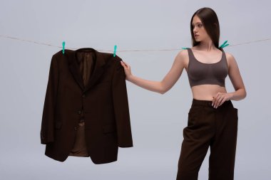 stylish teenage model in crop top pinned with clothespins on rope touching brown blazer hanging isolated on grey 