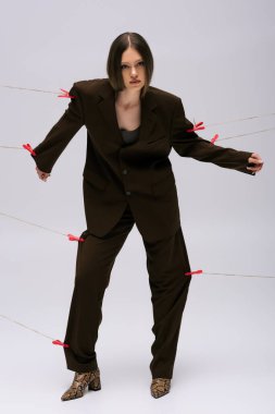 full length of stylish teenage girl in brown suit pinned with clothespins on rope posing on grey 