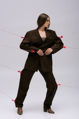 full length of teenage model in stylish brown suit pinned with clothespins on rope posing on grey 