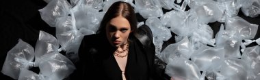 top view of brunette model in stylish suit posing around plastic bags on black, banner