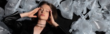 top view of brunette model with closed eyes lying around plastic bags on black, banner