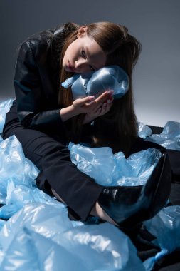 teenage model in stylish black suit posing around blue plastic bags on grey