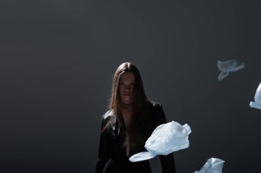 brunette model in stylish black suit posing near flying plastic bags on dark grey