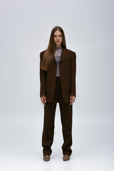full length of pretty teenage model in brown high-quality suit standing on grey