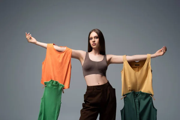 pretty model in crop top and trousers standing with outstretched hands and holding clothing on grey