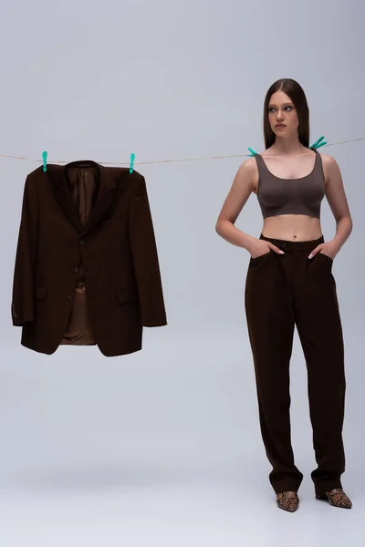 stylish teenage girl in crop top pinned with clothespins on rope near brown blazer hanging on grey