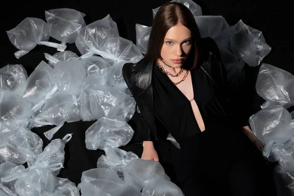 high angle view of pretty model in stylish suit posing around plastic waste on black