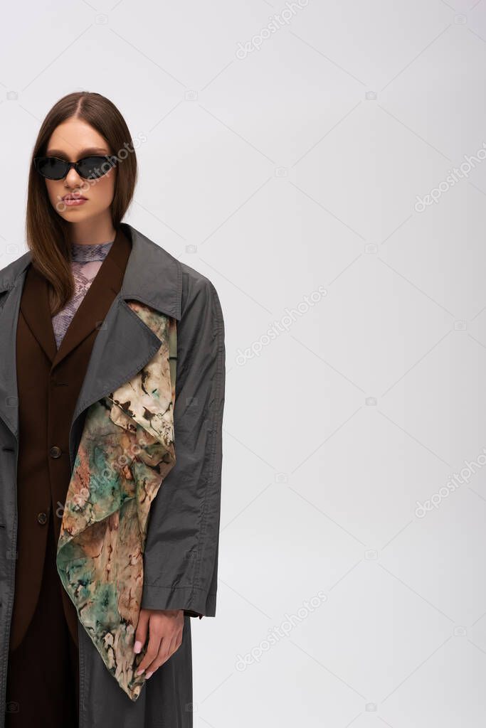 teenage model in stylish sunglasses and trench coat isolated on grey