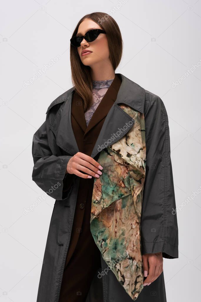low angle view of teenage model in stylish sunglasses and trendy trench coat isolated on grey