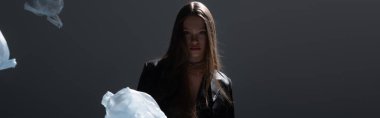 brunette model in stylish black suit posing near flying plastic bags on dark grey, banner