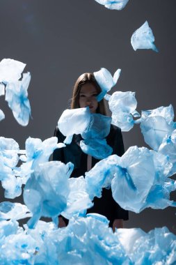 brunette model in suit posing near flying blue plastic bags on dark grey