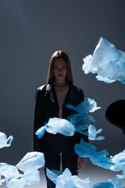 brunette model in black suit posing near flying blue plastic bags on dark grey