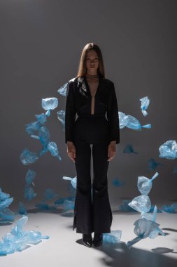 full length of pretty model in black suit posing near blue plastic bags on grey