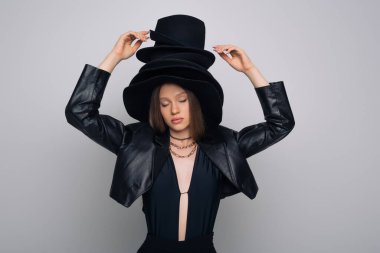 brunette model with closed eyes touching black hat on head isolated on grey