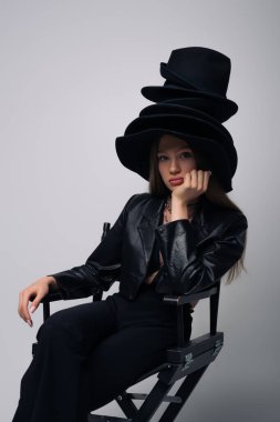 brunette model in leather jacket and different black hats sitting on chair isolated on grey