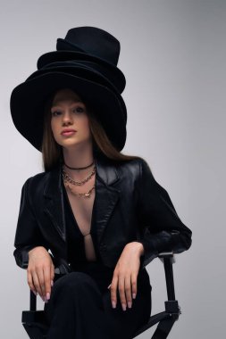 brunette model in leather jacket and different black hats sitting isolated on grey