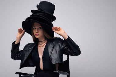 teenage model in leather jacket and different black hats sitting isolated on grey