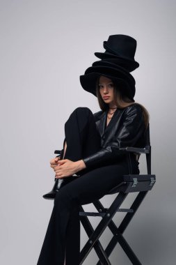 brunette model in black leather jacket and different black hats sitting on chair on grey