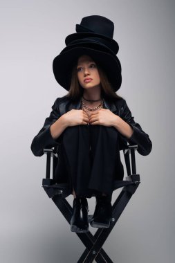 model in black leather jacket and different black hats sitting on chair on gray