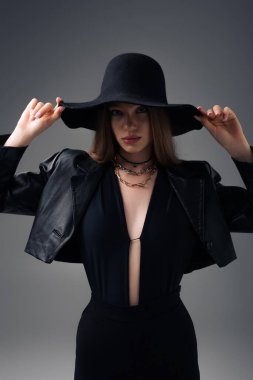 teenage model in leather jacket adjusting floppy hat isolated on grey