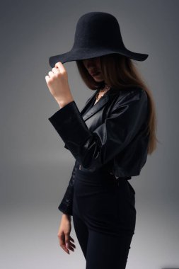 brunette teenage model in black leather jacket adjusting floppy hat isolated on grey