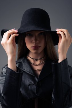 teenage model in black leather jacket adjusting floppy hat isolated on grey