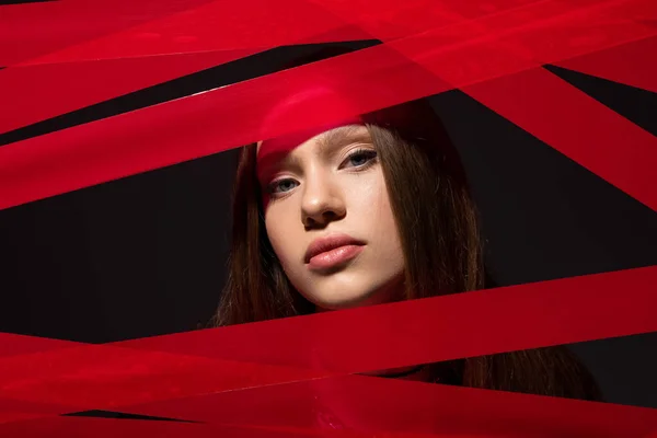 teenage model looking at camera through red strips from duct tape isolated on black, recycle fashion concept