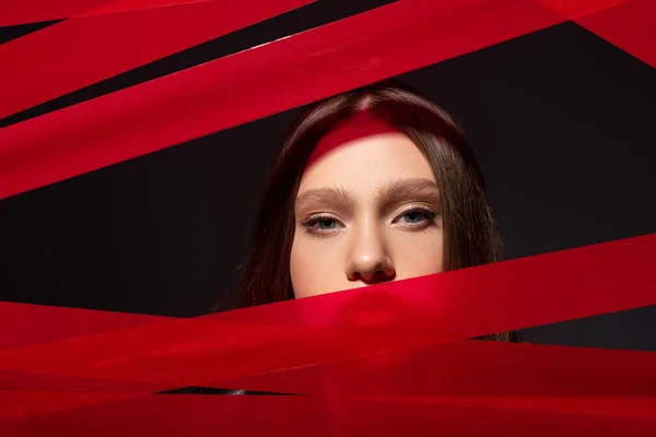teenage model looking at camera through red strips from adhesive tape isolated on black, recycle fashion concept