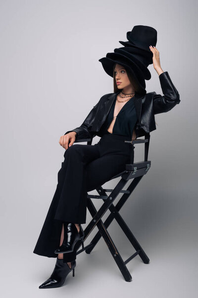 модель брюнетки в кожаной куртке и различных черных шляпах, сидящих на стуле на сером