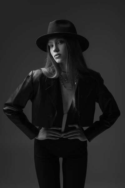 Монохромное фото модели-подростка в шляпе-шляпе, позирующей руками на бёдрах в студии 