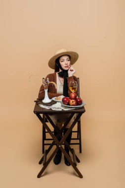 Stylish woman posing near wine and food on table on beige background 