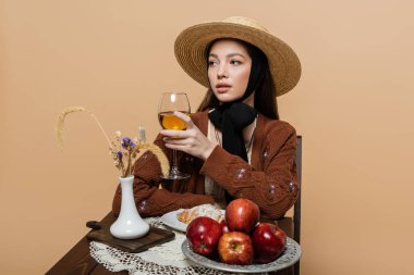 Stylish woman in sun hat holding wine near food on table isolated on beige 