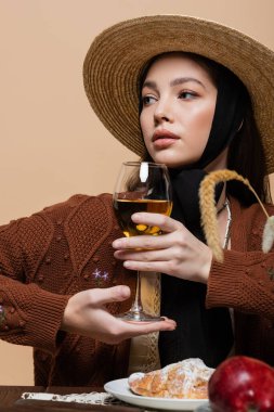 Stylish woman in straw hat posing with glass of wine near croissant on table isolated on beige 