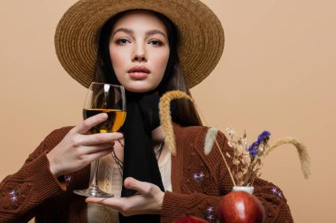 Portrait of stylish woman in sun hat holding wine near plants isolated on beige 
